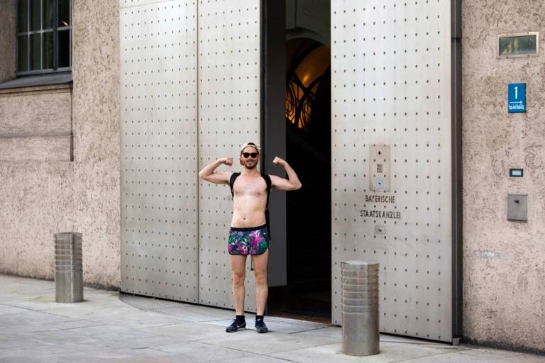 A man who is wearing swimming trunks is standing in front of the entrance of the Bayerische Staatskanzlei in Munich.