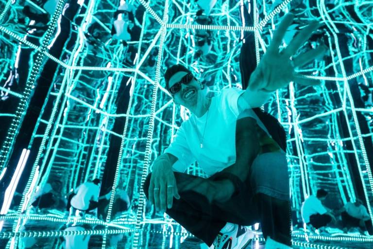 A man with sunglasses and a white T-shirt kneels in a blue-lit exhibition space that plays with illusions.