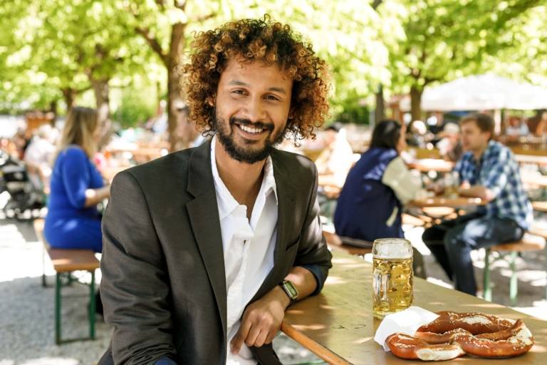 A man with curly hair and business jacket is sitting on a bench in a beer garden in Munich. On the table besides him is mug of beer and a pretzel.