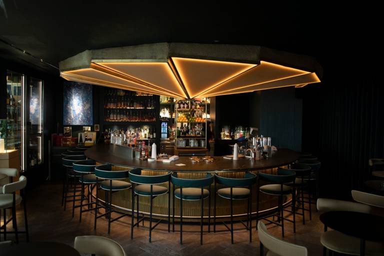 Stylish circular counter with bar stools, in background shelves with drinks, oranges, lemons ,limes and umbrella shaped lighting above the counter in hotel bar Ory in Munich.