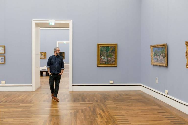 Un uomo con gli occhiali e la barba attraversa la Neue Pinakothek di Monaco di Baviera e guarda i dipinti.