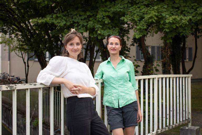 Two women lean against a bridge railing and smile into the camera.